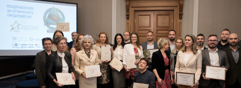 Large group of award recipients posing together at an awards ceremony in a hall, many holding certificates and trophies, in front of a screen reading "Congratulations for the high achievements in innovations for 2022!"